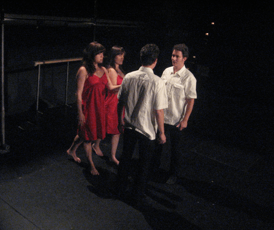 Vier Personen auf einer dunklen Bühne. Zwei Männer, die offensichtlich Zwillinge sind, stehen sich gegenüber und schauen sich an. Zwei Frauen, beide im roten Kleid, kommen von der Seite auf sie zu. Alles sieht wie gespiegelt aus.