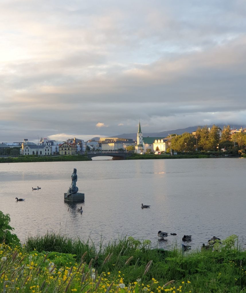 Da siehst Du einen Teich, mit Enten drauf. Im Wasser steht eine Statue (Mädchen, Nixe) und auf der gegenüberliegenden Seite sind Häuser und eine Kirche. Alles ist in warme Abendsonne getaucht.