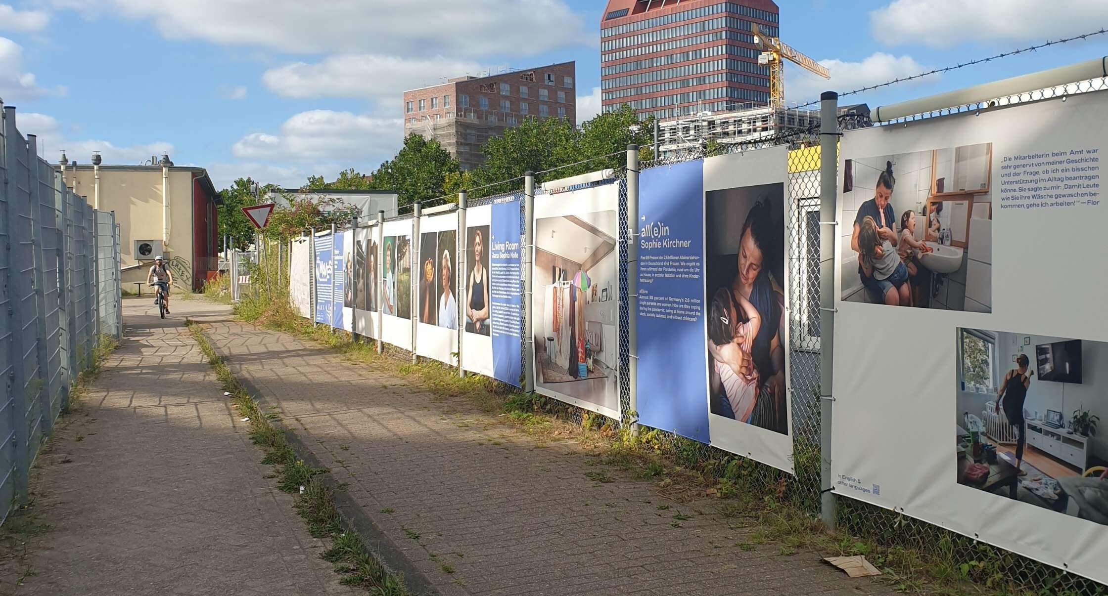 Da ist ein Weg und rechts vom Weg ein Zaun, an dem große Plakate mit Fotos und Texten aufgehängt sind.