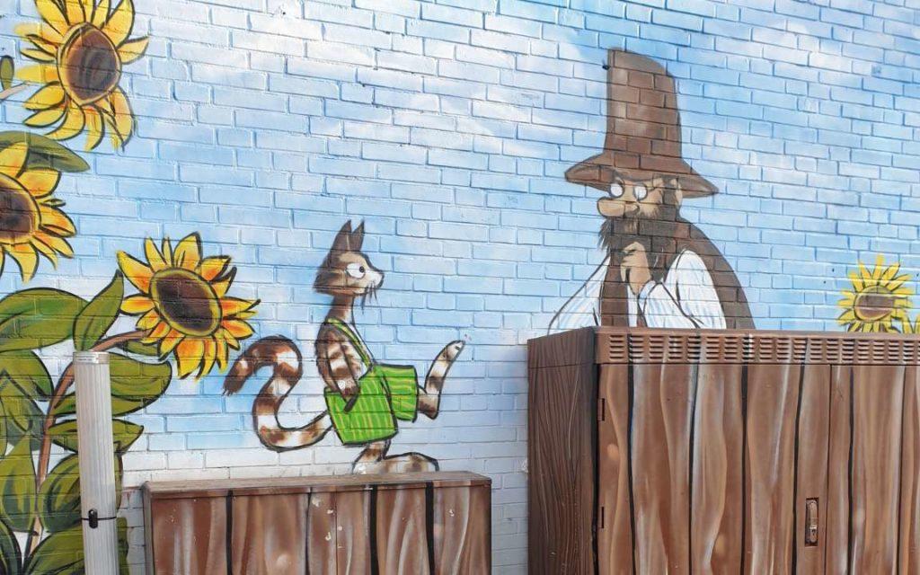 Eine bemalte Wand. Sie zeigt Petterson und Findus mit Sonnenblumen im Garten.