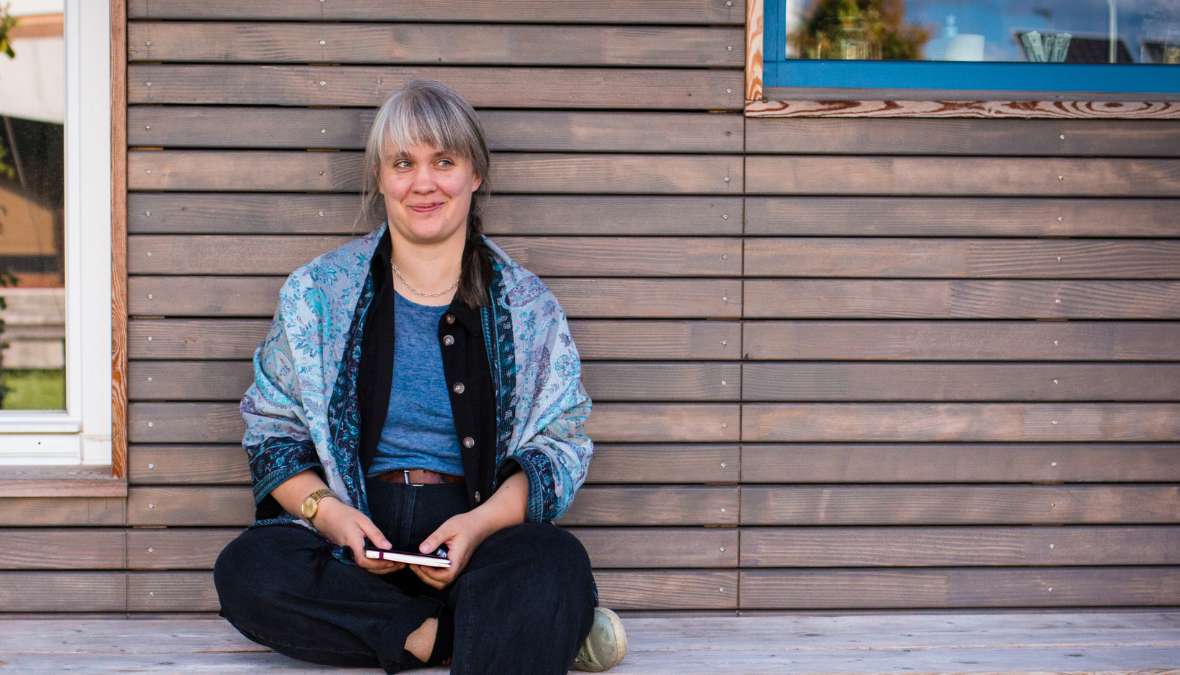 Paula Marie Berdrow by Ingrid Hagenhenrich, 2022 Paula sitzt vor einer Holzwand, hat ein Notizbuch in den Händen und schaut schmunzelnd, zur Seite.