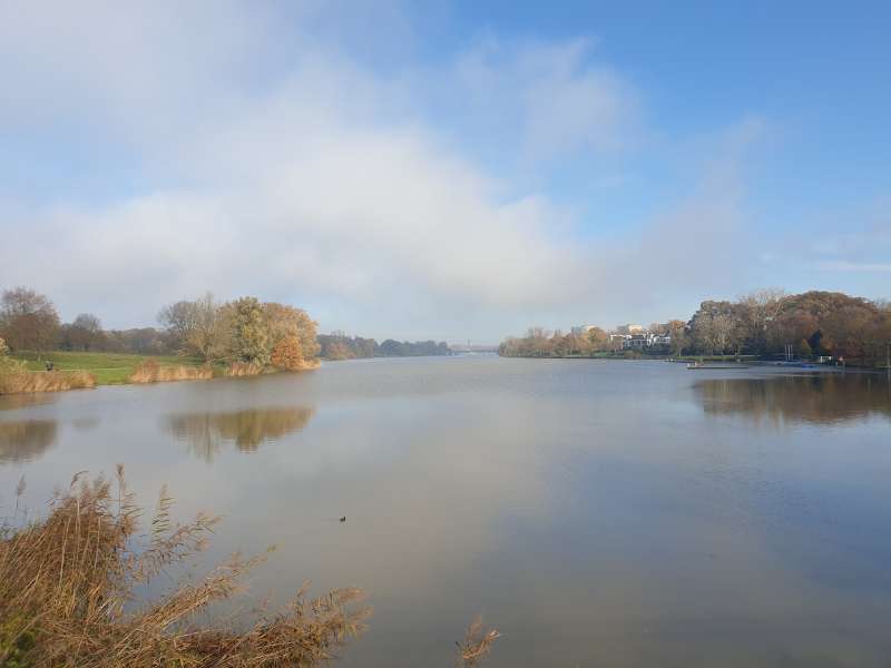 Ein großer See, von herbstlich braunen Bäumen gesäumt. Am blauen Himmel sind zur ein paar fluffige Wolken.