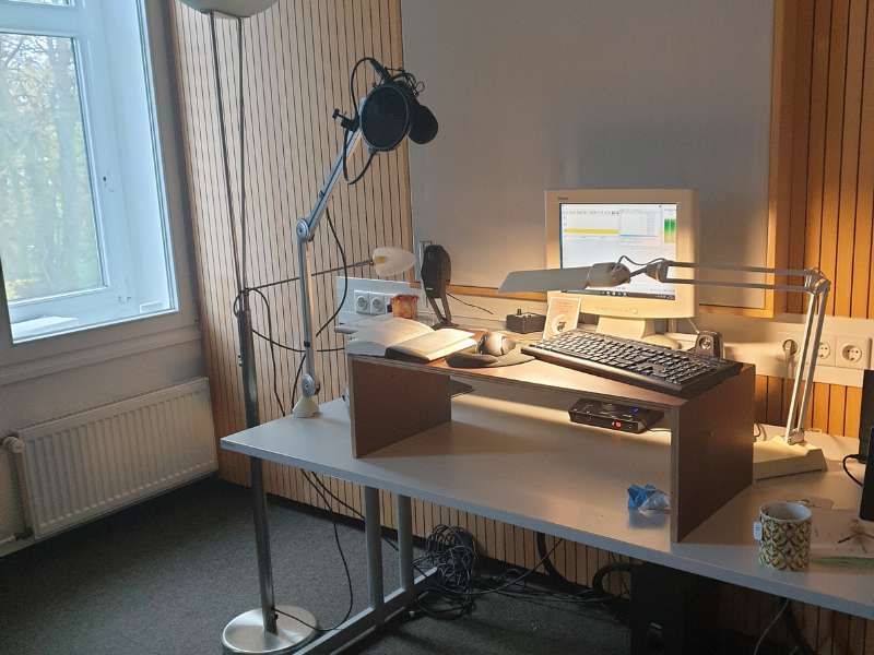 Ein Tonstudio mit einem Schreibtisch, Competer, Mikrofon und einem einfachen Aufsatz, auf dem Tastatur, Maus und ein Buch erhöht liegen.