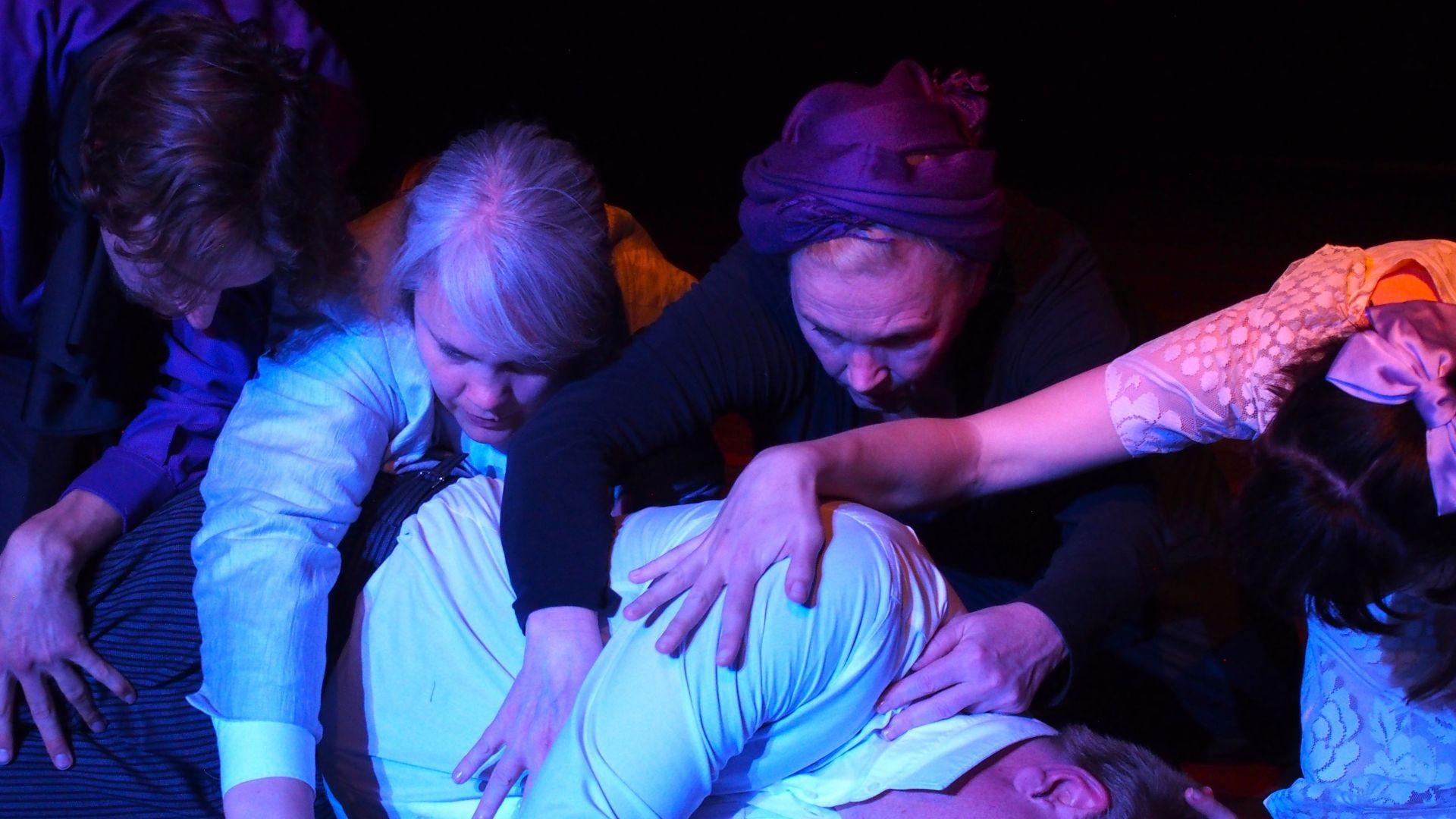 Schöne Fremde Theater en face 2024 Eine Szene auf der Bühne in bläulichem Licht. Eine Person liegt am Boden, die anderen beugen sich darüber und ihre Hände wandern über den liegenden Körper.