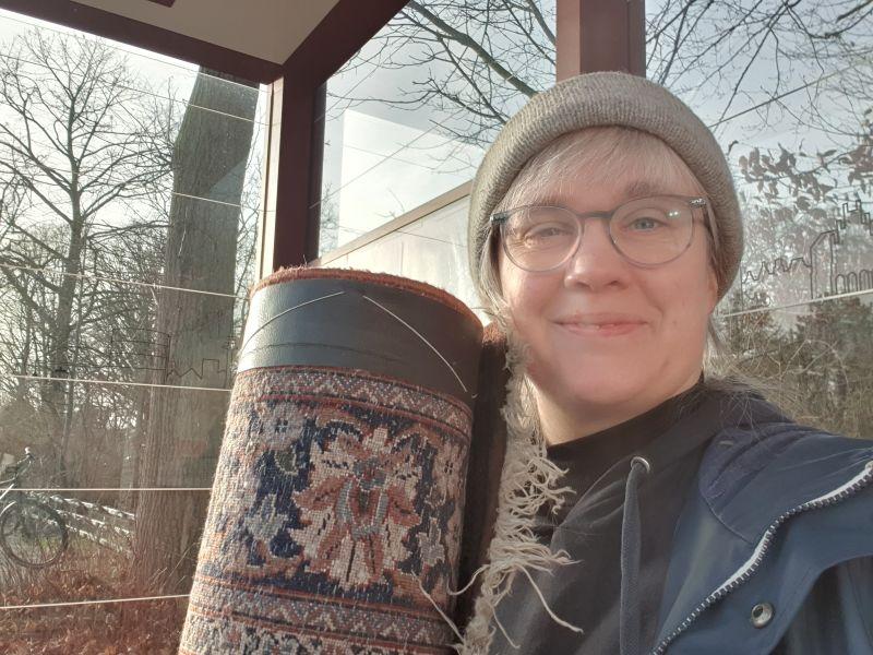 Paula mit einem aufgerollten Teppich an einer Bushaltestelle.