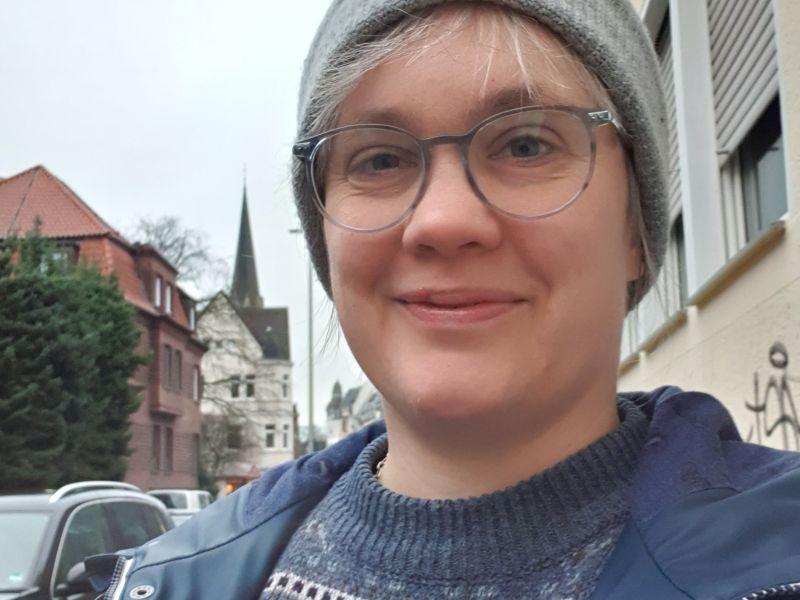 Paula, mit Mütze und Wollpullover, vor einer winterlichen Straße.