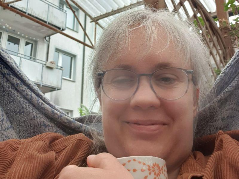 Paula mit einer Tasse Tee und beschlagenen Brillengläsern in einem Hängesitz.