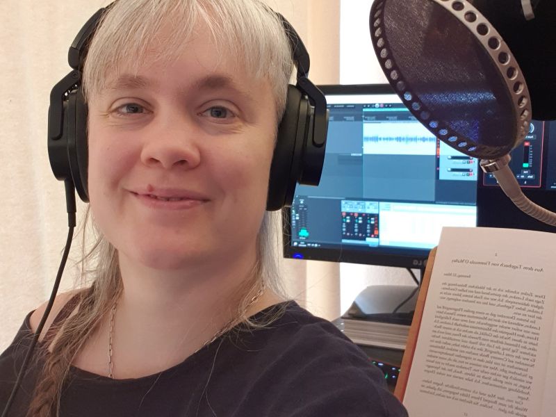 Paula mit Kopfhörern. Im Hintergrund ein Buch und ein Bildschirm.