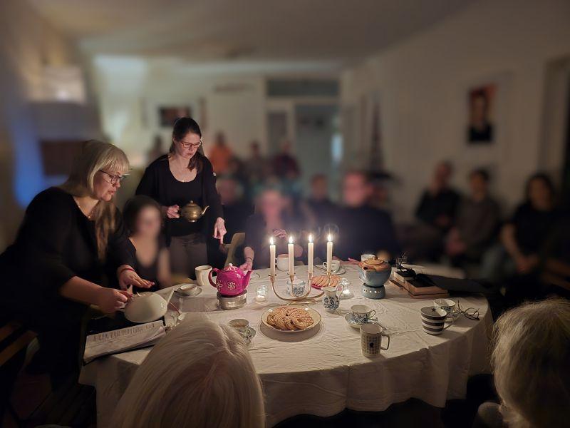 Paula und Petra schenken Tee ein, an einer großen Tafel bei Kerzenlicht. Am Tisch und im Hintergrund sind unscharf weitere Menschen zu erkennen.