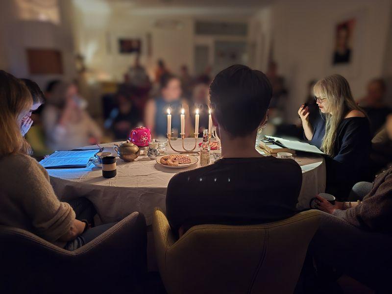 Ein Foto von einer Lesung. Paula und ihre Kollegin sitzen sich an einem gedeckten Tisch mit Kerzen und Gebäck gegenüber. Paula liest etwas vor. Ihre Hand macht eine gruselige Geste.