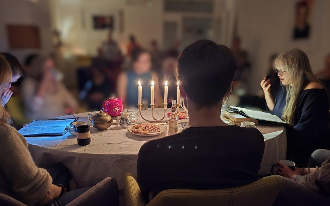 Paula sitzt an einem Tisch, mit vielen anderen Leuten zusammen, bei Kerzenlicht, und liest mit einer dramatischen Geste vor.