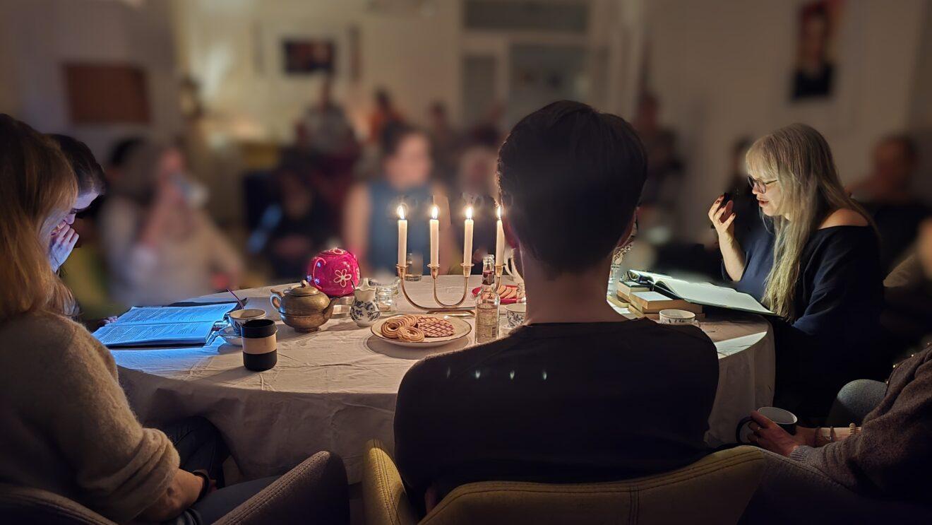 Paula sitzt an einem Tisch, mit vielen anderen Leuten zusammen, bei Kerzenlicht, und liest mit einer dramatischen Geste vor.
