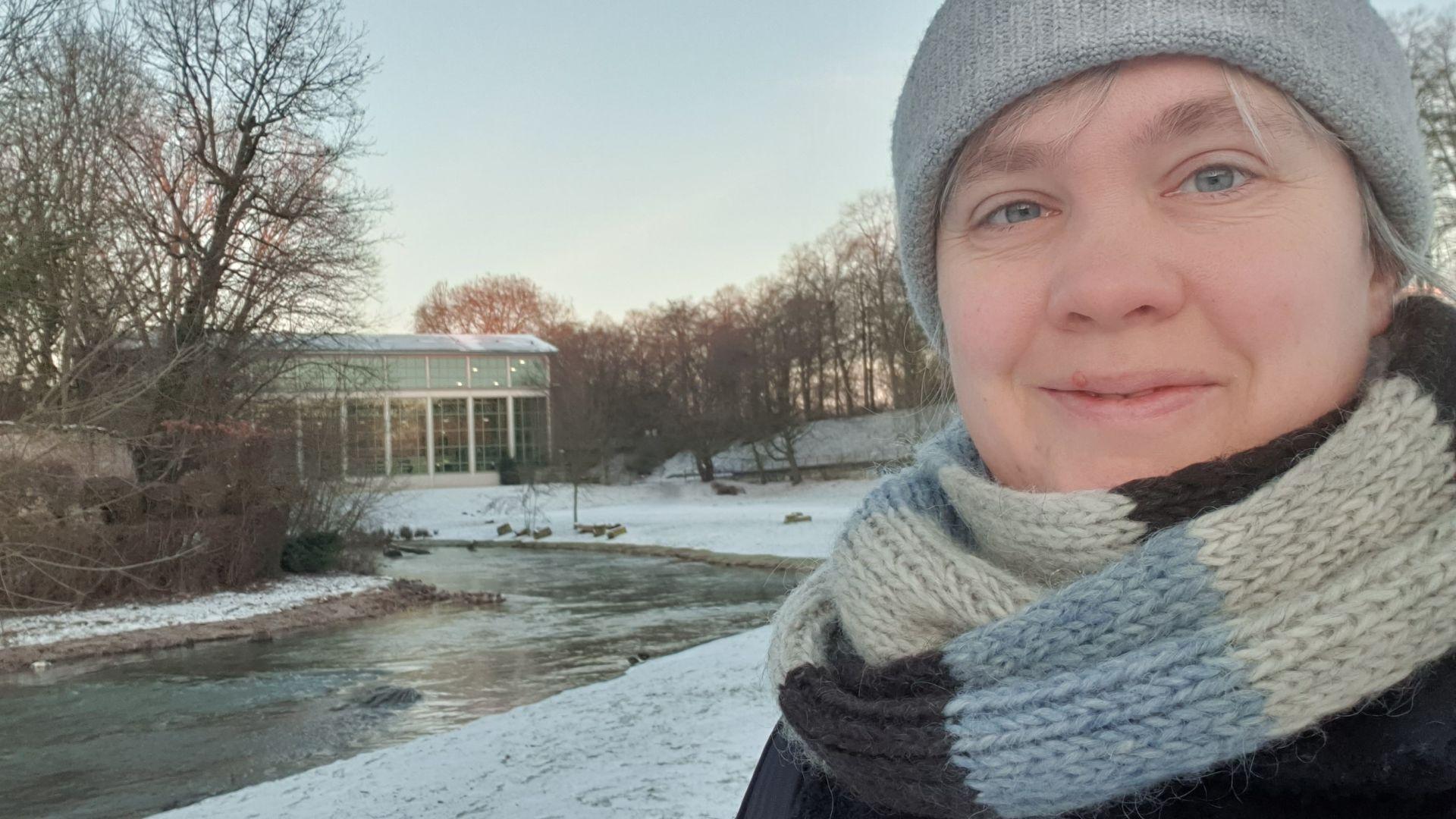 Meine Ziele bis zum 31. März Paula in Mütze und Schal, im Hintergrund ein winterlicher Fluss und ein Glasgebäude.