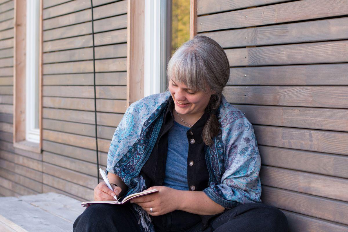 Paula Marie Berdrow by Ingrid Hagenhenrich, 2025 – Schreiben Paula sitzt draußen vor einem Holzhaus und schreibt in ein Notizbuch.