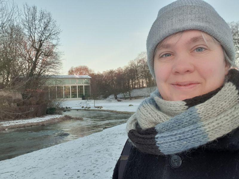 Paula in Mütze und Schal, im Hintergrund ein winterlicher Fluss und ein Glasgebäude.