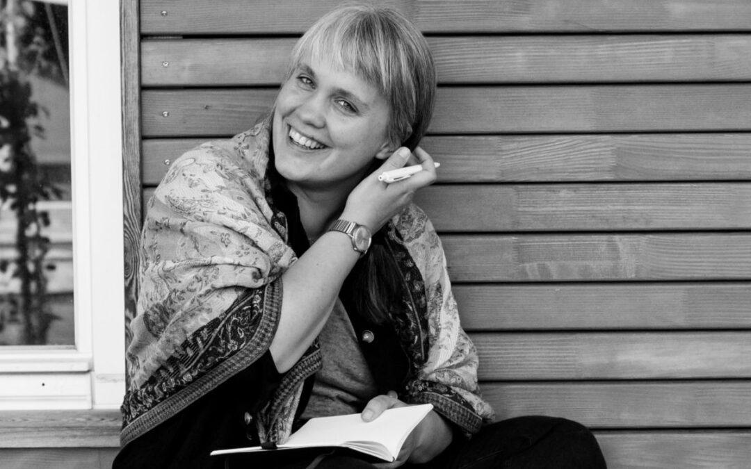 Paula sitzt vor einer hölzernen Wand mit einem Notizbuch. Sie schaut lachend in die Kamera und streicht sich die Haare hinters Ohr.