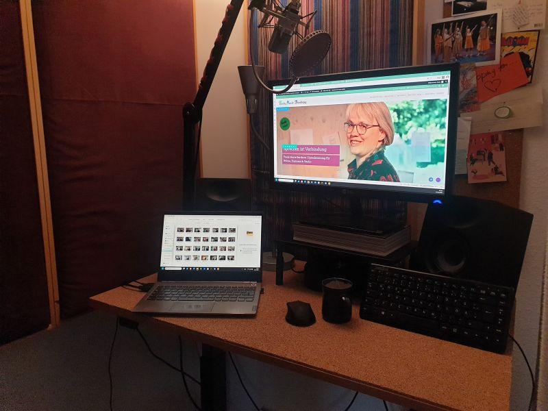 Blick in Paulas Studio: Ein Schreibtisch mit Korkauflage, Akustikpaneele an den Wänden. Der Monitor zeigt Paulas Website, und auf dem Tisch steht noch zusätzlich ein Laptop.
