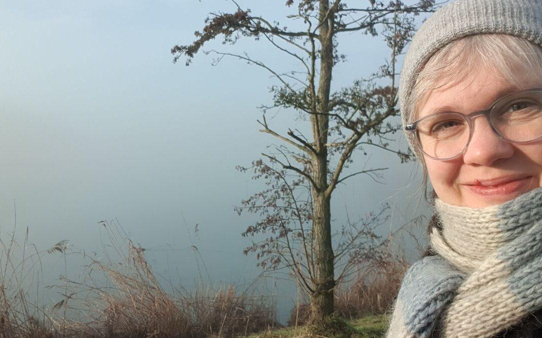 Paula mit Mütze und Schal, im hintergrund ist ein Baum und ein Seeufer, das im Nebel verschwimmt.
