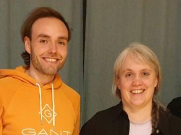 Website Startbild 1904x591 px (1200 x 400 px) - 1 Paula steht in einem Raum mit großen Fenstern. Ihre Hände schweben vor dem Körper nach oben. Rechts im Bild sieht man den Arm einer weiteren Person nach oben schweben.