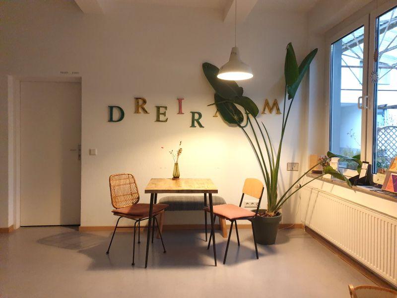 Ein Tisch mit zwei Stühlen, über den Tisch steht in großen Buchstaben an der Wand: "Dreiraum".
