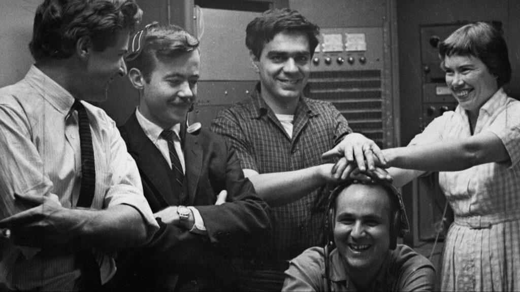 Drei Männer und eine Frau (Pauline Oliveros) stehen in einem Tonstudio und legen die Hände auf den Kopf eines vierten, sitzenden Mannes.