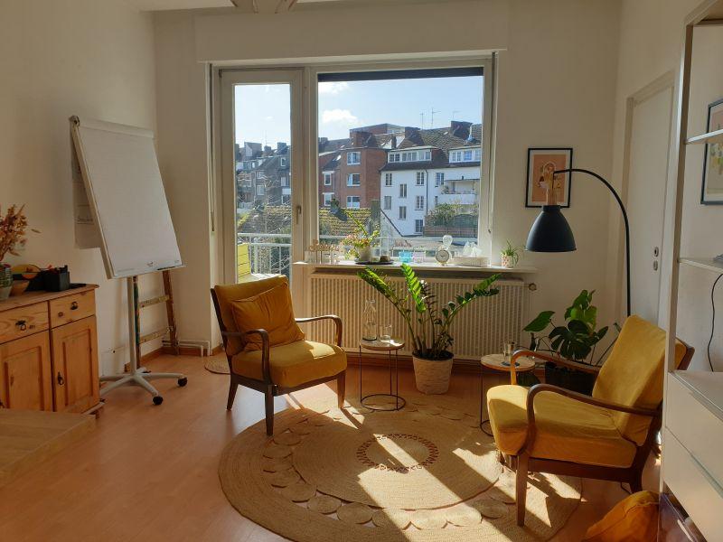 Ein warm erleuchteter Raum mit zwei Sesseln, Pflanzen und einem Fenster + Tür zum Balkon.