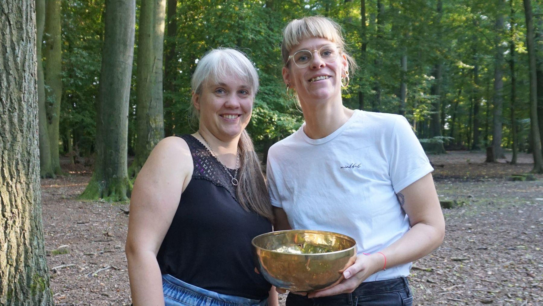Paula und Laura stehen nebeneinander unter Bäumen. Laura hat eine große messingfarbene Klangschale in der Hand.